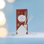 A festive Apple Cinnamon diffuser featuring a charming white gingerbread house vessel, styled in a snowy setting with soft glowing lights.