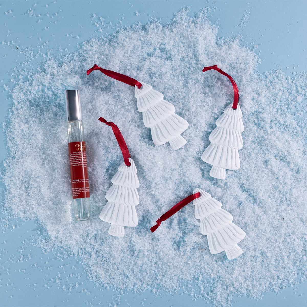 A set of four white Christmas tree hanging diffusers with red ribbons, styled alongside a festive fragrance spray on a snowy blue backdrop.