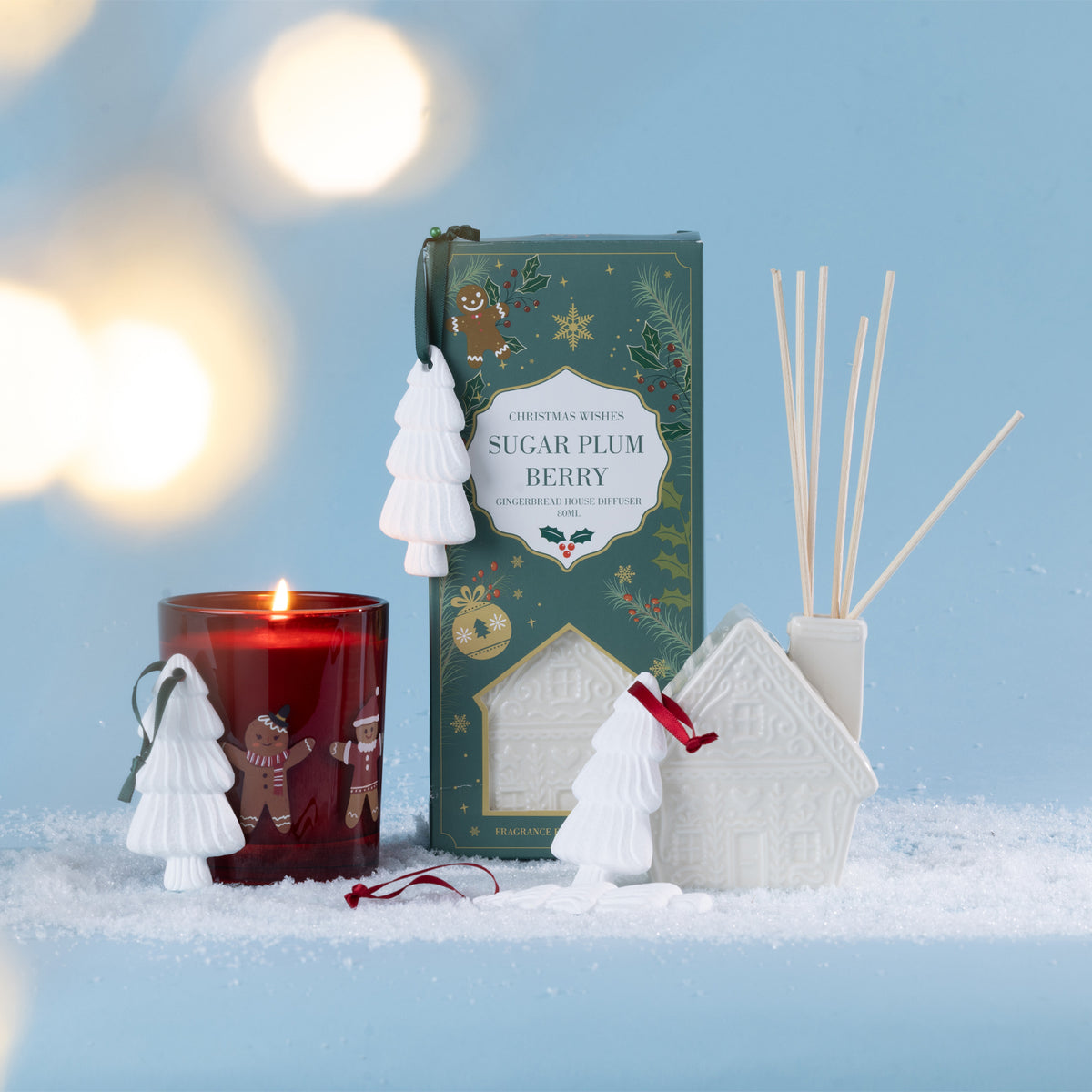 A festive fragrance collection featuring a gingerbread house diffuser and coordinating candle, styled with white Christmas tree ornaments in a snowy setting.