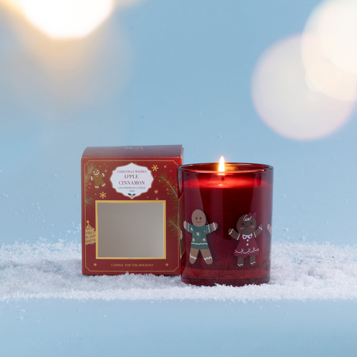 A festive Apple Cinnamon candle in a rich red glass vessel decorated with playful gingerbread characters, styled in a snowy setting with soft glowing lights.