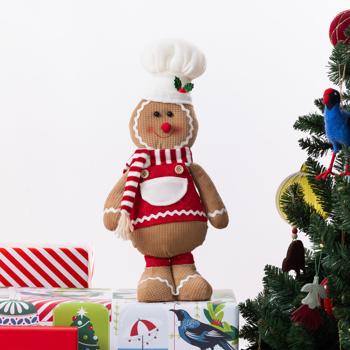 A gingerbread Christmas figurine dressed as a chef, wearing a white hat with green holly accents, a red and white striped scarf, and red dress, standing on top of wrapped gifts next to a decorated Christmas tree.