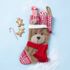 A plush teddy bear Christmas stocking with a red and white plaid pattern, a red knit scarf, and a red-and-white plaid Santa hat, styled on a light blue background with candy canes and gingerbread men.