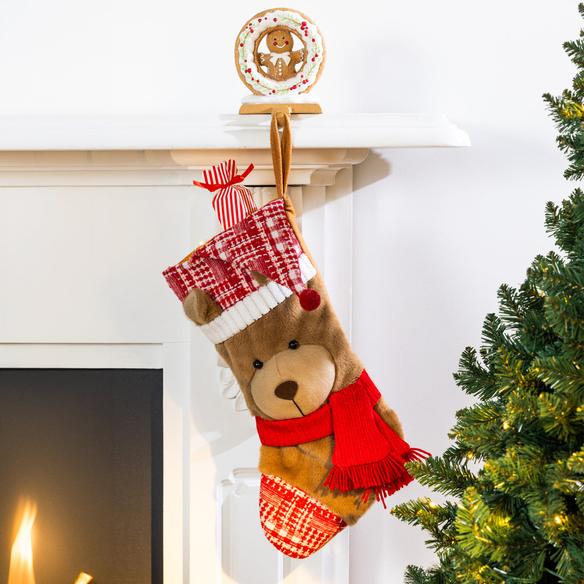 A plush teddy bear Christmas stocking with a plaid Santa hat and red scarf, hanging from a white fireplace mantel next to a lit Christmas tree.