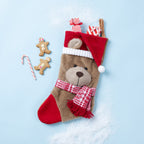 A plush teddy bear Christmas stocking with a red Santa hat, red-and-white plaid scarf, and red toe, styled on a light blue background with two gingerbread cookies and a candy cane.