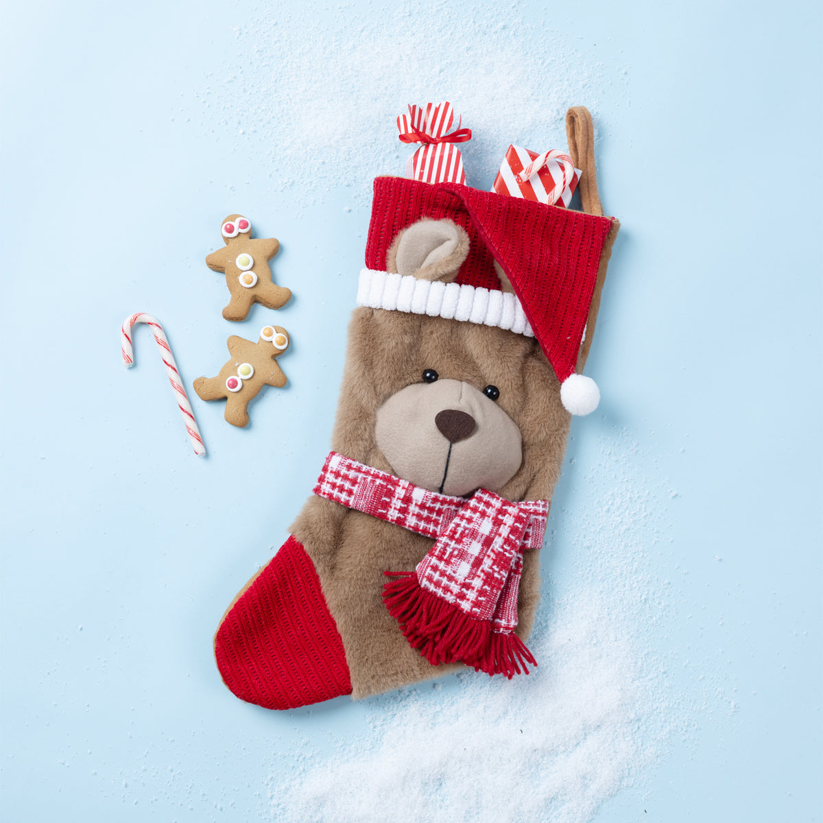 A plush teddy bear Christmas stocking with a red Santa hat, red-and-white plaid scarf, and red toe, styled on a light blue background with two gingerbread cookies and a candy cane.