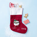 A red and white Christmas stocking with a snowman face and the words 'LET IT SNOW' on the front, styled on a light blue background with candy canes, gingerbread cookies, and wrapped gifts.