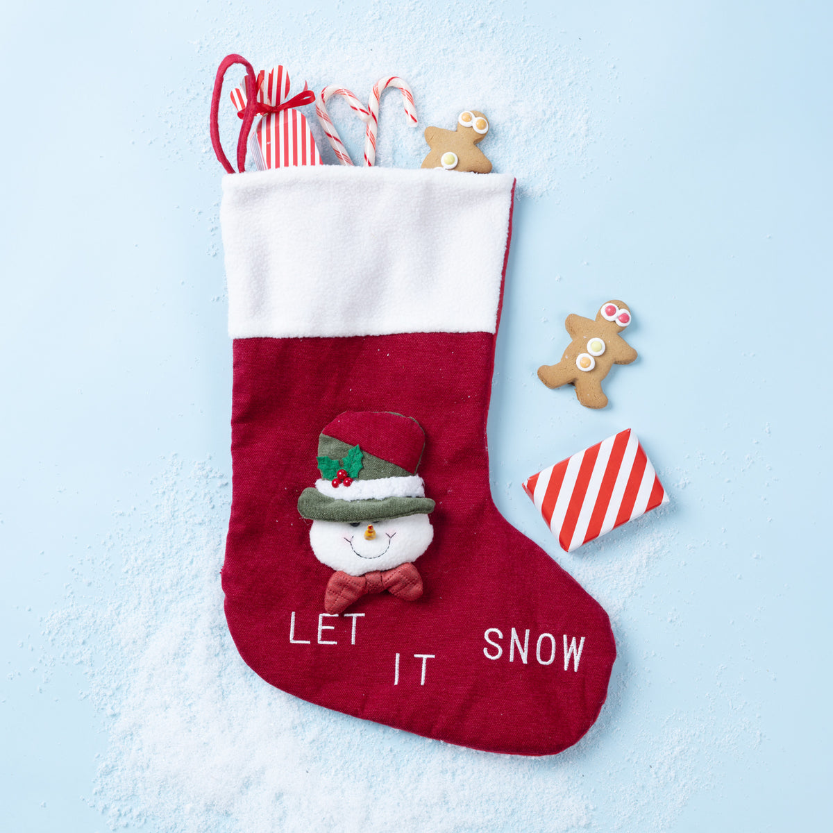 A red and white Christmas stocking with a snowman face and the words 'LET IT SNOW' on the front, styled on a light blue background with candy canes, gingerbread cookies, and wrapped gifts.