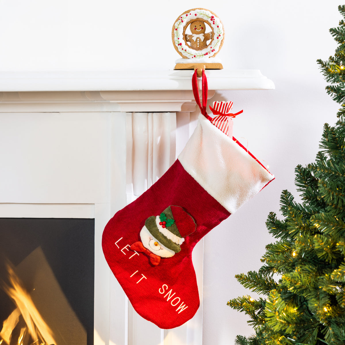 A red Christmas stocking with a white cuff hangs from a mantel. The stocking features an embroidered snowman and the words 'LET IT SNOW'.