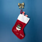 A red Christmas stocking with a white cuff, featuring a snowman decoration and the embroidered text 'LET IT SNOW'. It hangs from a gold bow ornament against a solid blue background.