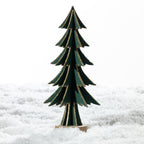 A small decorative Christmas tree made of layered dark green wood with gold glitter edges, standing on a round wooden base in a snowy white setting.