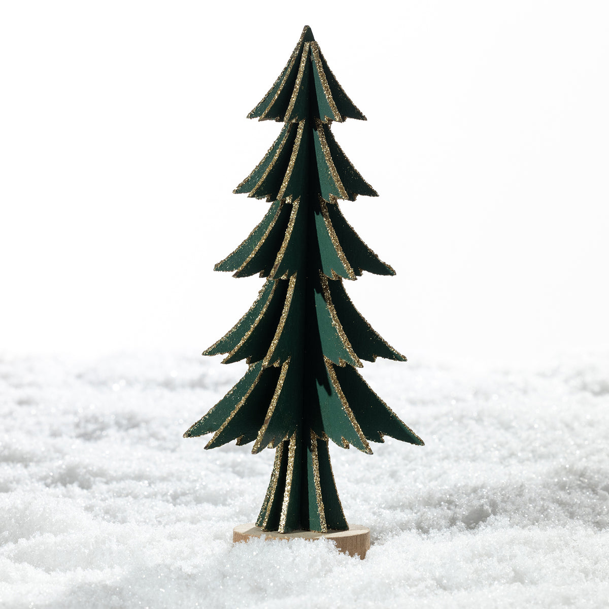 A small decorative Christmas tree made of layered dark green wood with gold glitter edges, standing on a round wooden base in a snowy white setting.