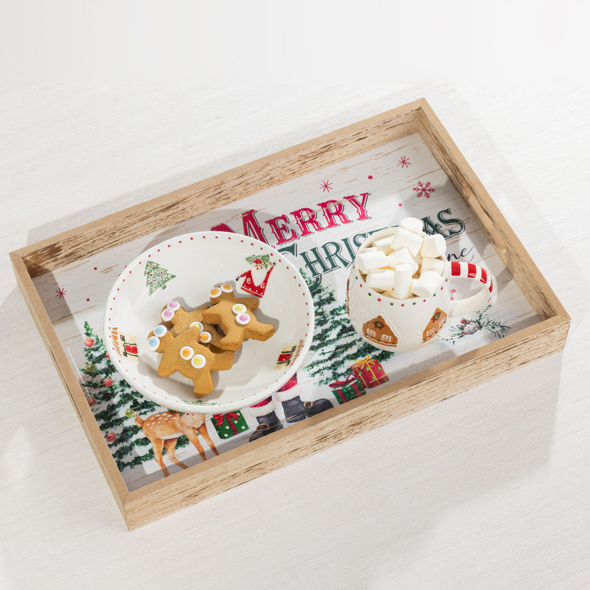 A rectangular wooden serving tray with a printed Christmas scene and the words “Merry Christmas,” styled with a white plate of gingerbread cookies and a white mug filled with marshmallows on a light surface.