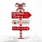 A festive Christmas-style directional signpost with three red and white signs reading “Gingerbread Bakery,” “Hot Cocoa,” and “North pole,” topped with a red-and-white striped bow, shown standing in snow against a white background.
