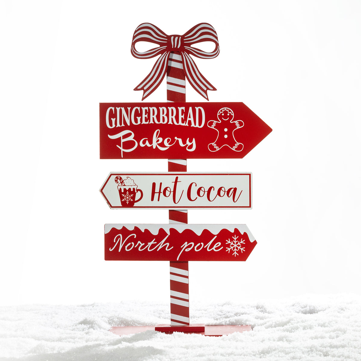 A festive Christmas-style directional signpost with three red and white signs reading “Gingerbread Bakery,” “Hot Cocoa,” and “North pole,” topped with a red-and-white striped bow, shown standing in snow against a white background.