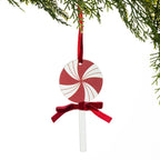 A peppermint lollipop Christmas ornament hanging from a green pine branch. The ornament features a round candy head with a red and white spiral pattern, a red velvet ribbon bow at the base, and a clear plastic stick.