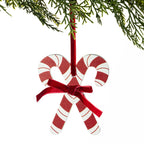 A Christmas ornament shaped like two candy canes crossed at the bottom, with a red ribbon bow at the center and a red hanging loop. The ornament is shown against a white background with pine branches at the top.