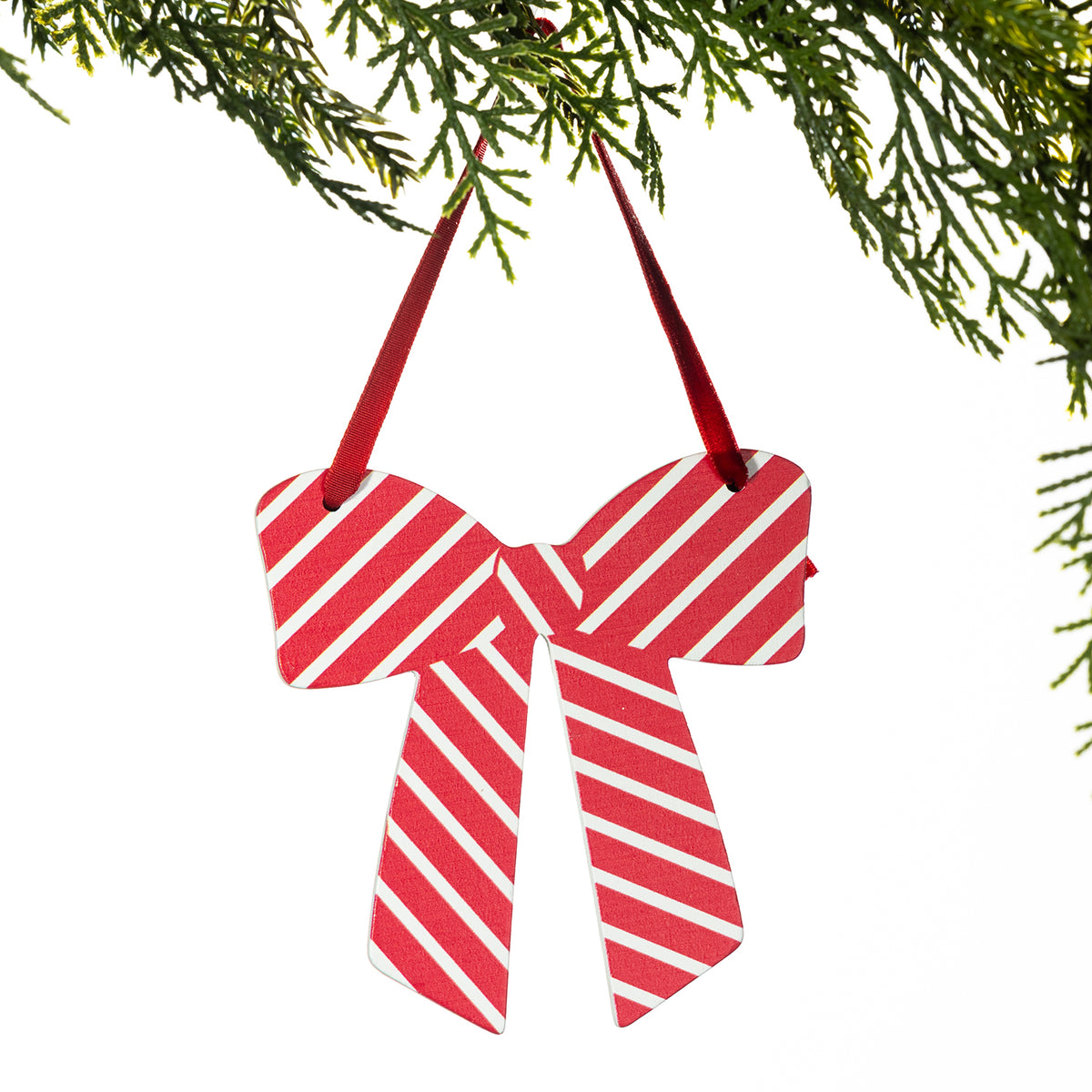 A red and white striped bow-shaped Christmas ornament hanging from a green pine branch against a white background.