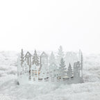 Christmas Wishes Metal Village Candle Holder – White metal village-style tealight holder featuring layered cut-out houses, trees and festive woodland details.