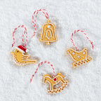 Four gingerbread holiday ornaments shaped like a bird in a Santa hat, a bell, a sleigh, and a rocking horse, arranged on a white textured background. Each ornament is decorated with white icing and hangs from a red and white striped string.