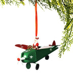 A Christmas ornament shaped like a green airplane with Santa Claus sitting in the cockpit, hanging from an orange ribbon against a white background with pine branches framing the top right.