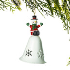 A white metal Christmas bell ornament with snowflake cutouts, topped by a small snowman figure in a red coat, green scarf, and black hat, hanging from a green pine branch against a white background.