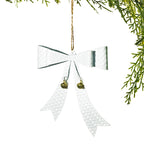 A silver hammered metal Christmas ornament shaped like a bow with two small gold bells and a jute hanging string, shown against a white background with green pine branches at the top right.