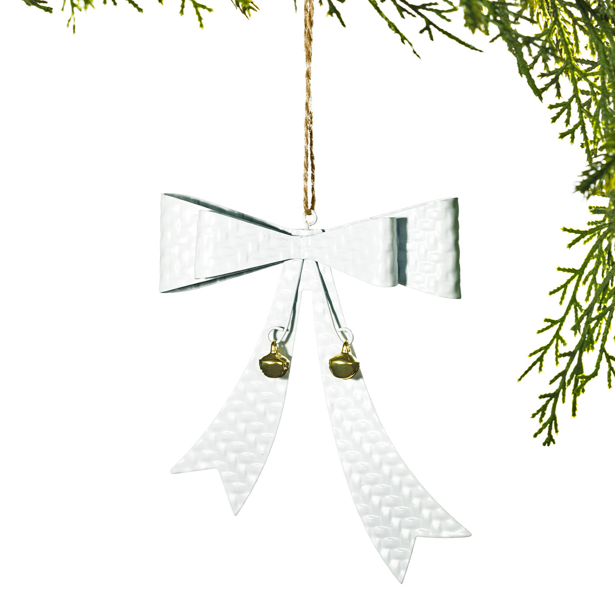 A silver hammered metal Christmas ornament shaped like a bow with two small gold bells and a jute hanging string, shown against a white background with green pine branches at the top right.
