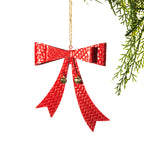 A red texture bow Christmas ornament with two small gold bells and a hanging string, shown against a white background with green pine branches in the corner.