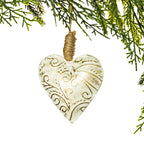 A heart-shaped wooden Christmas ornament with a distressed cream finish and carved scrollwork, hanging from a natural jute string wrapped around a pine branch, set against a solid white background.