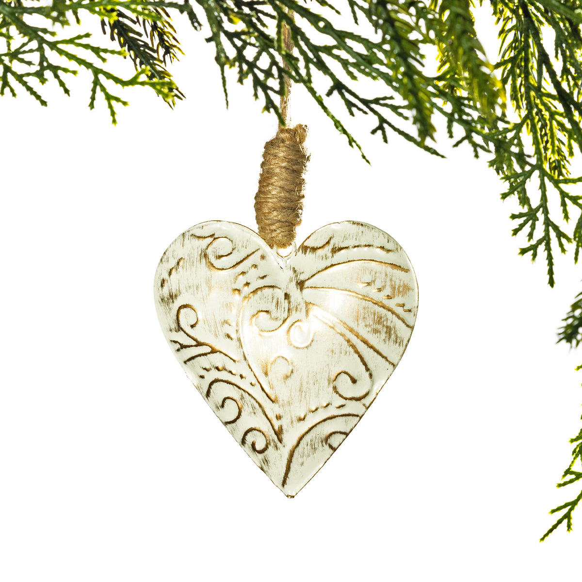 A heart-shaped wooden Christmas ornament with a distressed cream finish and carved scrollwork, hanging from a natural jute string wrapped around a pine branch, set against a solid white background.