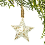 A distressed white ceramic star ornament with gold embossed scrollwork, hanging from a rustic jute twine loop against a white background with green pine branches framing the top and right edges.
