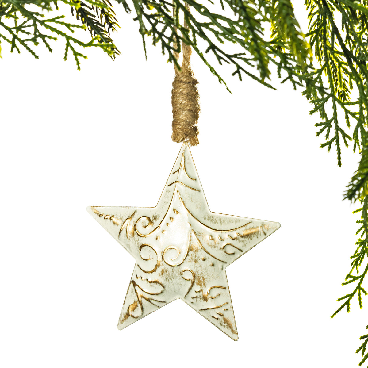 A distressed white ceramic star ornament with gold embossed scrollwork, hanging from a rustic jute twine loop against a white background with green pine branches framing the top and right edges.