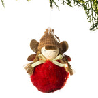 A festive Christmas angel ornament featuring a brown knit hat, gold glitter wings, a cream ribbon bow, and a fluffy red body, hanging against a white background with pine branches at the top.