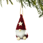 A small felt Christmas gnome ornament hanging from a tree branch against a white background. The gnome has a red pointed hat with a cream star, a white fluffy beard, and cream-colored feet.