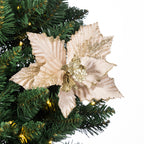 A large artificial poinsettia-style Christmas ornament in pale blush pink with gold glitter accents, shown attached to green artificial tree branches with warm white string lights against a white background.
