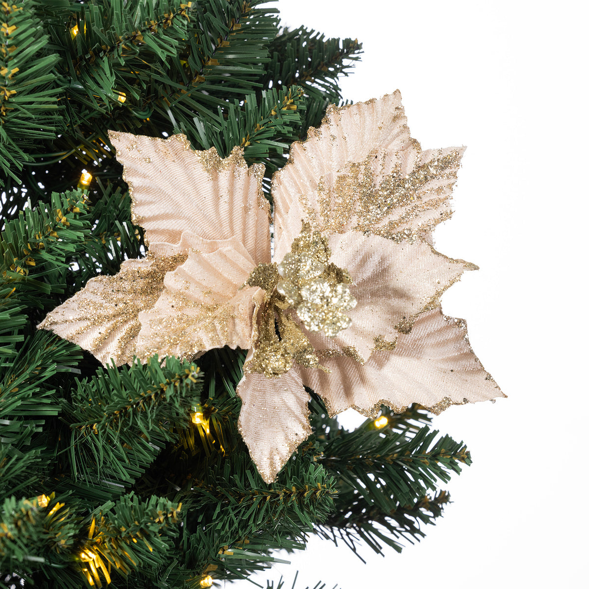 A large artificial poinsettia-style Christmas ornament in pale blush pink with gold glitter accents, shown attached to green artificial tree branches with warm white string lights against a white background.