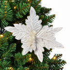 A white glitter poinsettia Christmas tree ornament hanging on a green artificial pine tree with warm yellow lights.