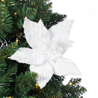 A white poinsettia artificial Christmas tree ornament with glittery textured petals and small rhinestone accents, shown hanging against a green artificial pine tree with warm string lights.