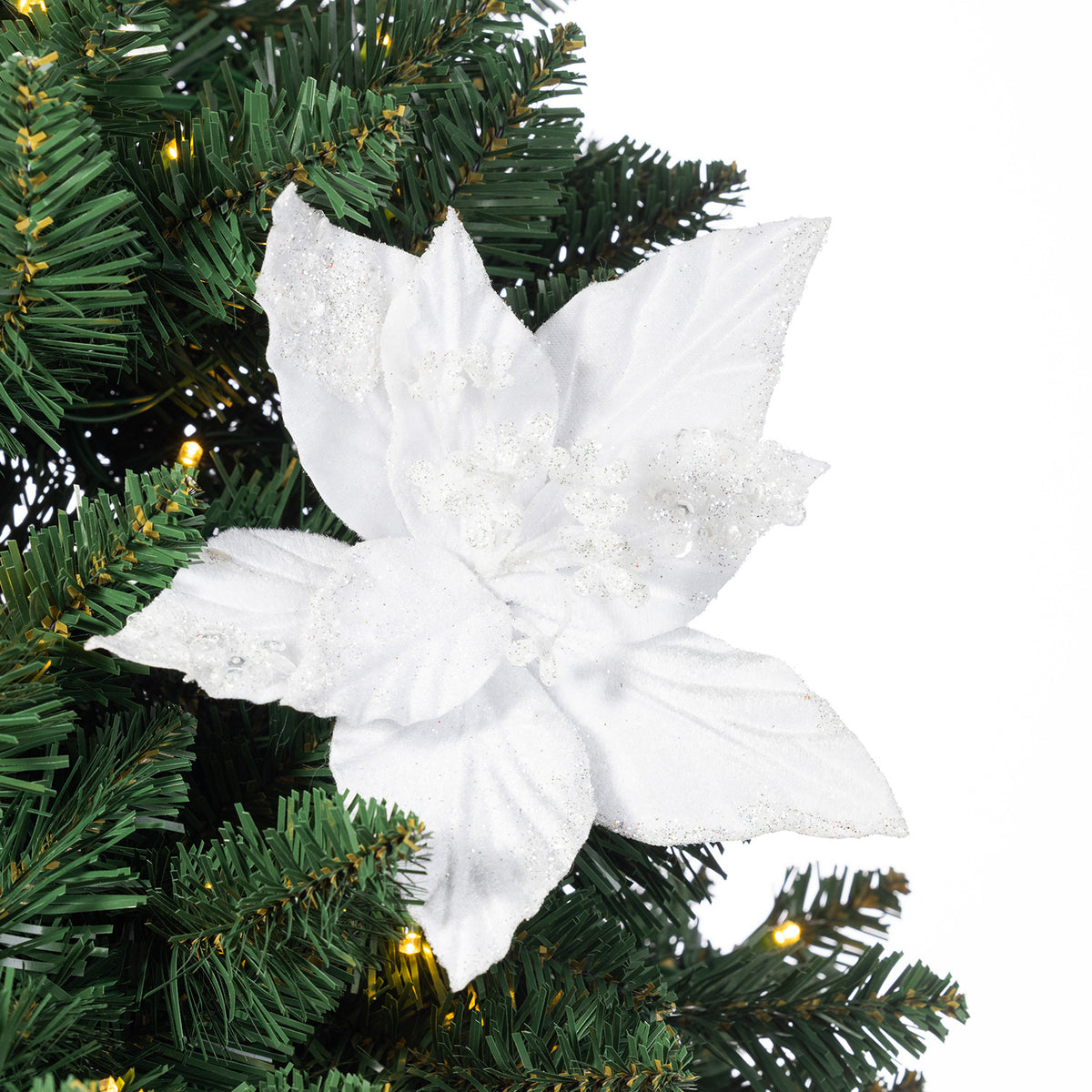 A white poinsettia artificial Christmas tree ornament with glittery textured petals and small rhinestone accents, shown hanging against a green artificial pine tree with warm string lights.