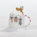 A white ceramic mug decorated with Christmas-themed hanging ornaments, filled with marshmallows and topped with a gingerbread man cookie, set against a clean white background with faux snow.