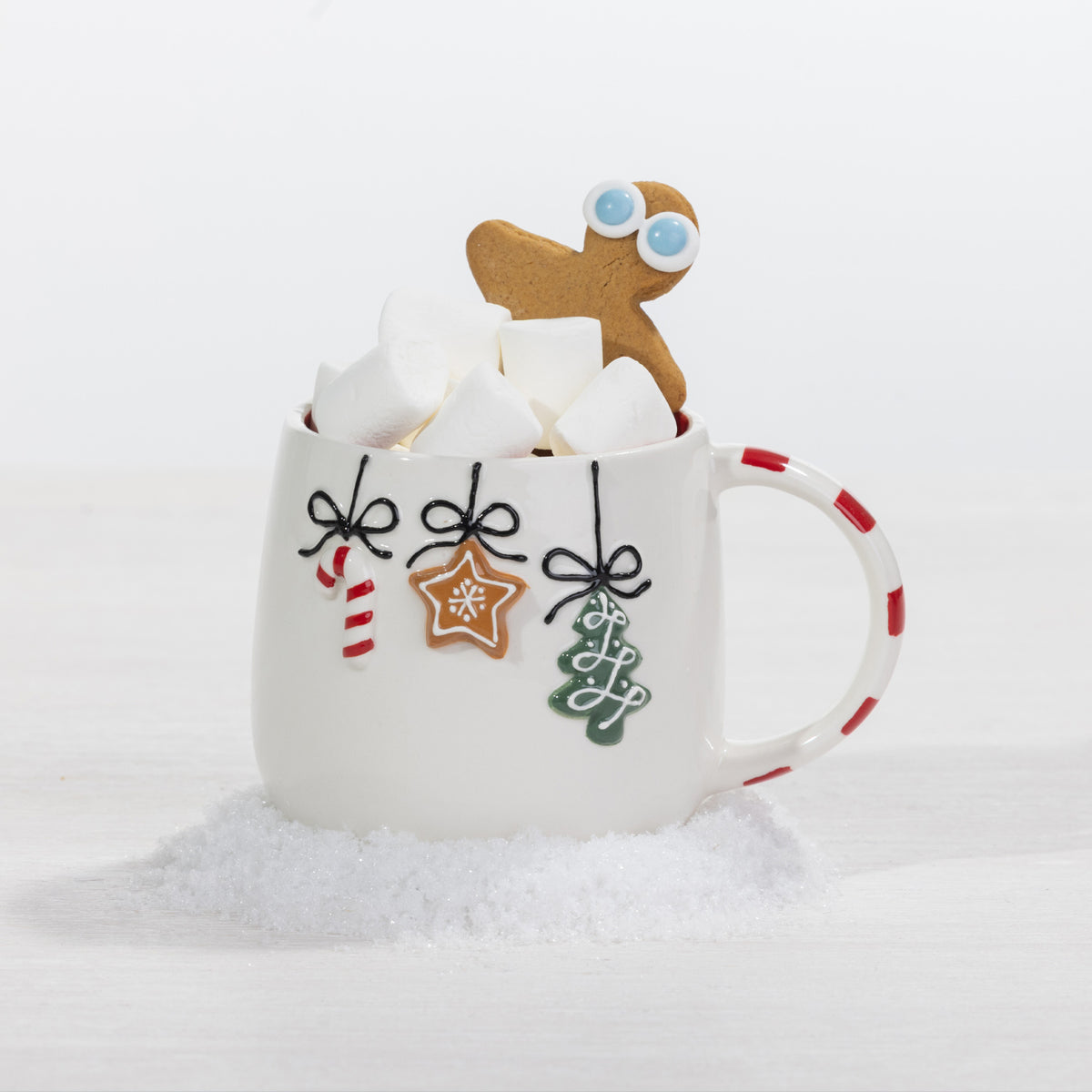 A white ceramic mug decorated with Christmas-themed hanging ornaments, filled with marshmallows and topped with a gingerbread man cookie, set against a clean white background with faux snow.