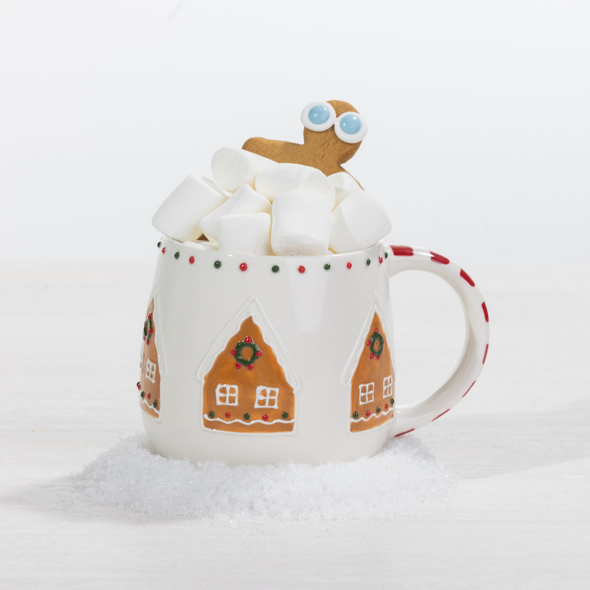A festive gingerbread house mug filled with marshmallows and topped with a playful gingerbread character, styled against a snowy white backdrop.