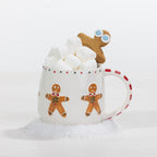 A white ceramic mug decorated with gingerbread men, filled with marshmallows and topped with a small gingerbread cookie, set against a clean white background with faux snow at the base.