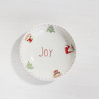 A white ceramic Christmas-themed bowl with the word 'Joy' written in red in the center, decorated with red dotted trim, green Christmas trees, and red-and-green Santa Claus illustrations around the rim.