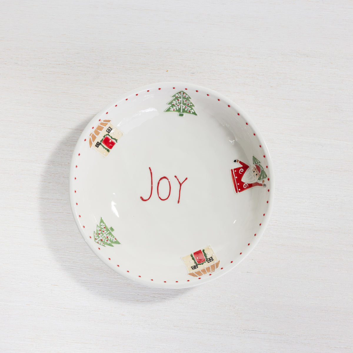 A white ceramic Christmas-themed bowl with the word 'Joy' written in red in the center, decorated with red dotted trim, green Christmas trees, and red-and-green Santa Claus illustrations around the rim.