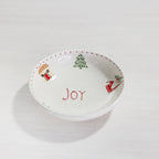 A white ceramic Christmas-themed dish or bowl with the word 'JOY' written in red in the center, decorated with a green Christmas tree and small holiday illustrations around the rim, set on a light wood surface.