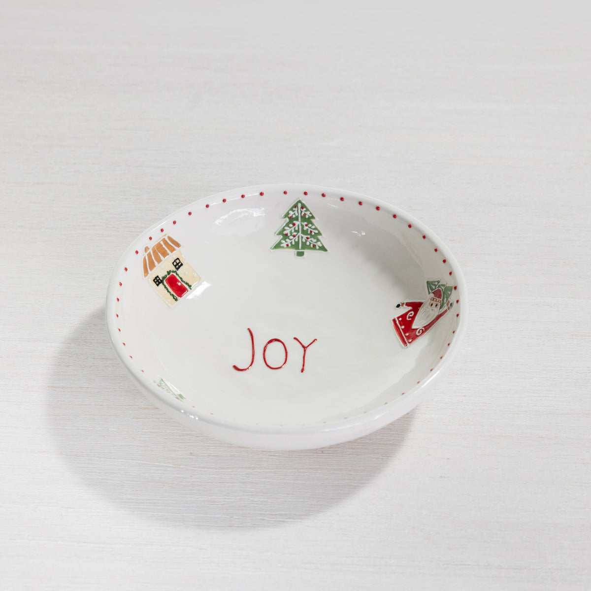 A white ceramic Christmas-themed dish or bowl with the word 'JOY' written in red in the center, decorated with a green Christmas tree and small holiday illustrations around the rim, set on a light wood surface.