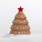A ceramic Christmas tree container or candle holder in a warm brown glaze, decorated with white icing-style swirls and snowflakes, topped with a red star, set against a clean white background with a light layer of faux snow at the base.
