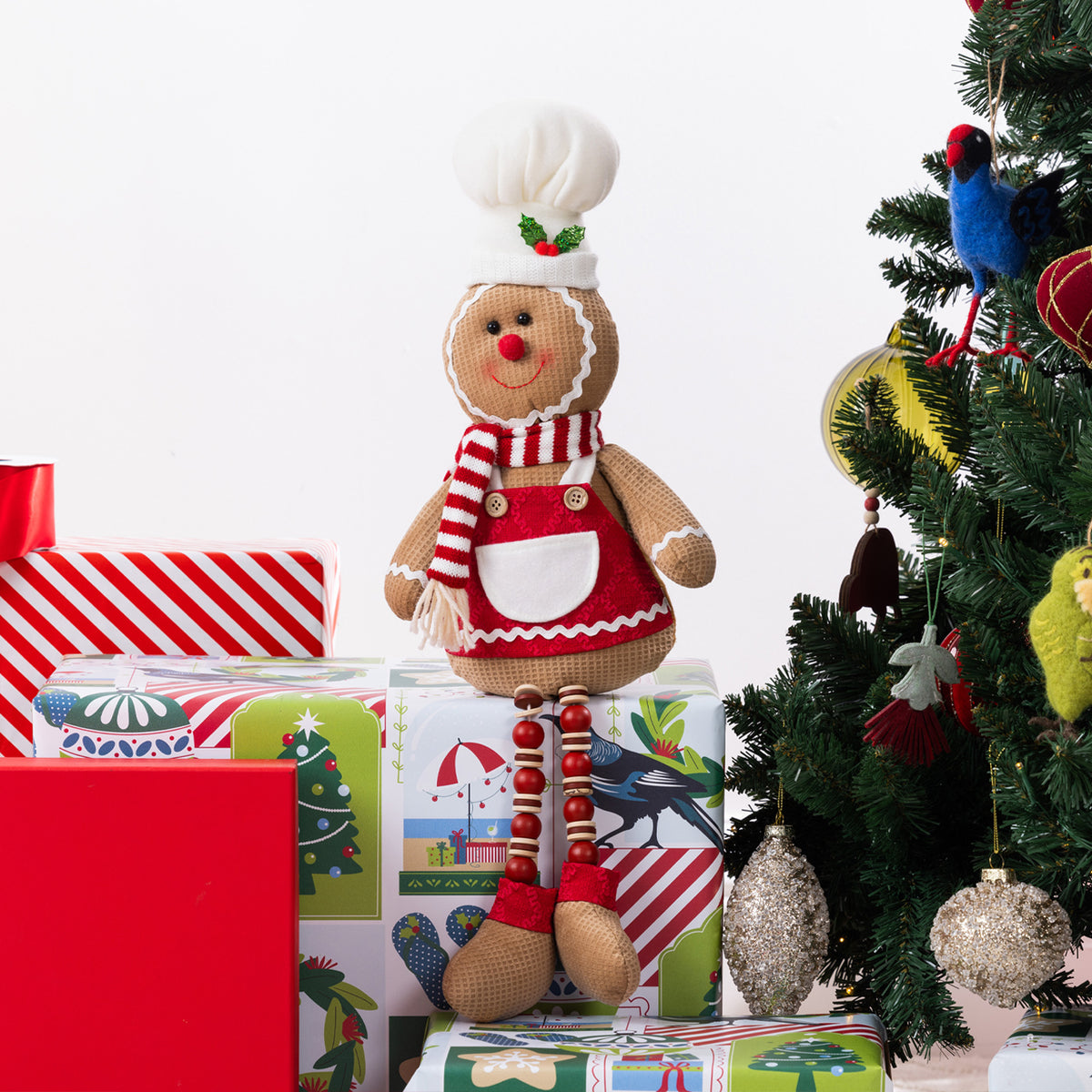 A gingerbread figurine sitting on Christmas presents and next to a Christmas Tree.
