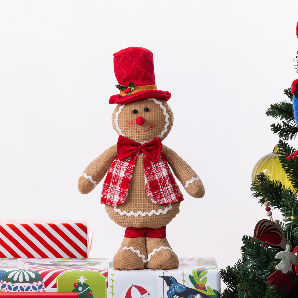 A gingerbread figurine standing on Christmas presents and next to a Christmas Tree.
